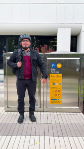 Man standing next to a bike locker while wearing a bicycle helmet and holding a backpack slung behind his shoulder