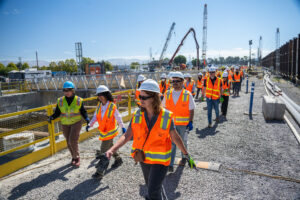 More than a dozen people wearing construction gear walking through a construction site